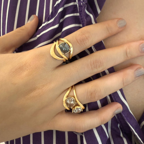 A close-up of a person's hand wearing two gold rings with stone accents, resting over a purple and white striped garment.