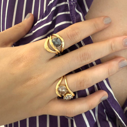 A close-up of a person's hand wearing two gold rings with stone accents, resting over a purple and white striped garment.