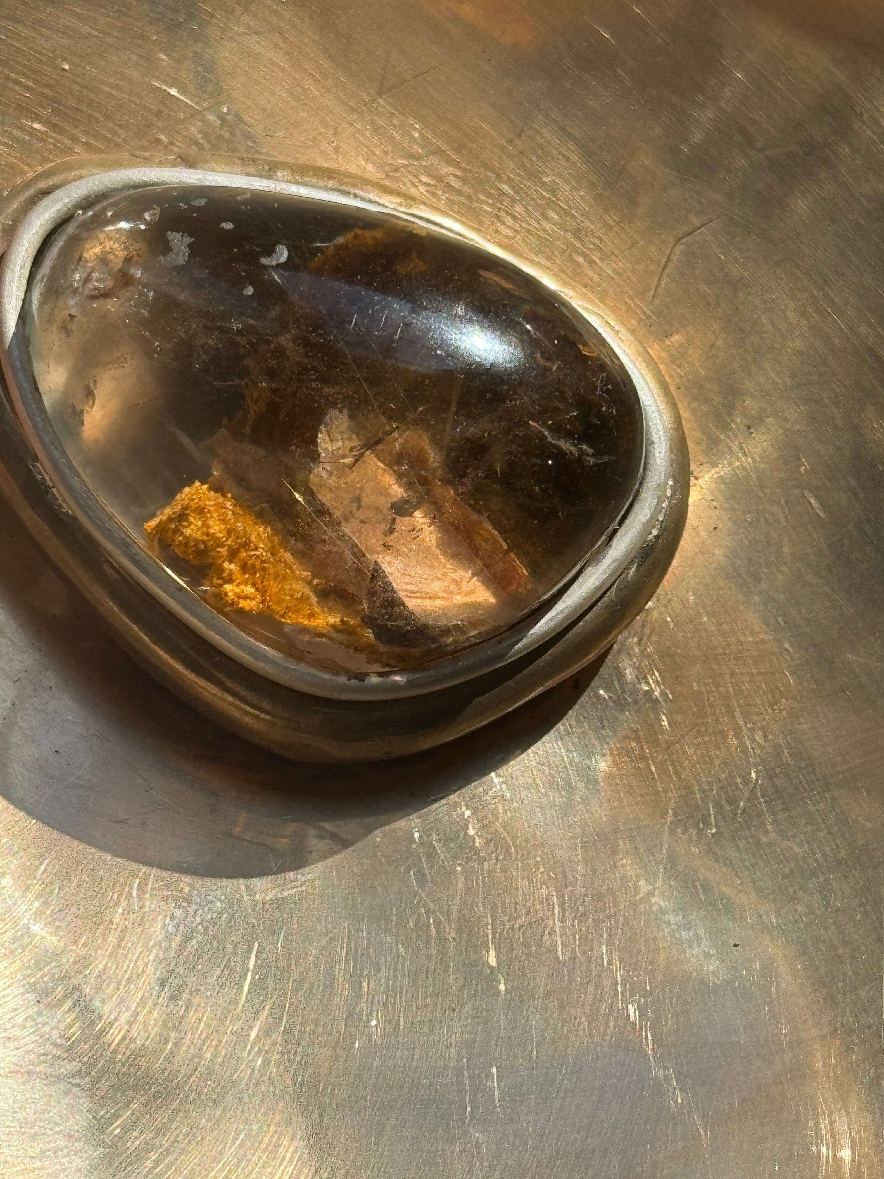 Brown quartz stone with gold inclusions in a silver bezel on a bronze metallic surface of a bowl 