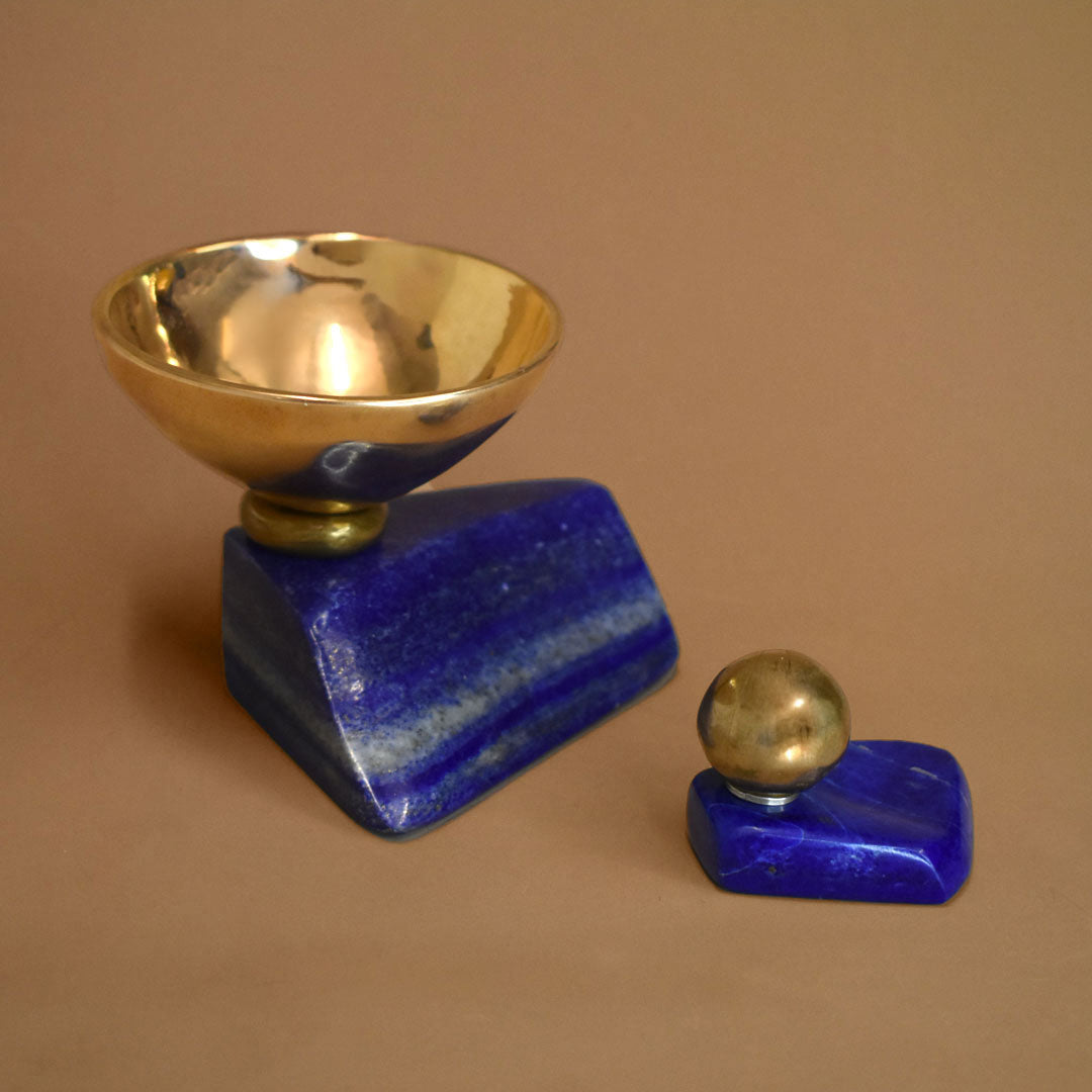 Bronze bowl adhered to a slab of lapis lazuli paired with a smaller piece of lapis lazuli with a small bronze ball adhered to it on a beige background