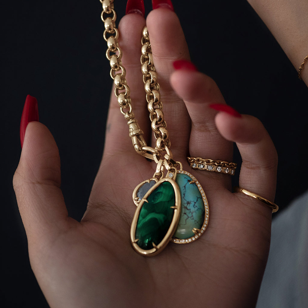 A hand with red nails holding a one of a kind chain with a malachite charm in a bezel setting with claw prongs set in yellow gold with white diamond accent stone with two other charms behind it.