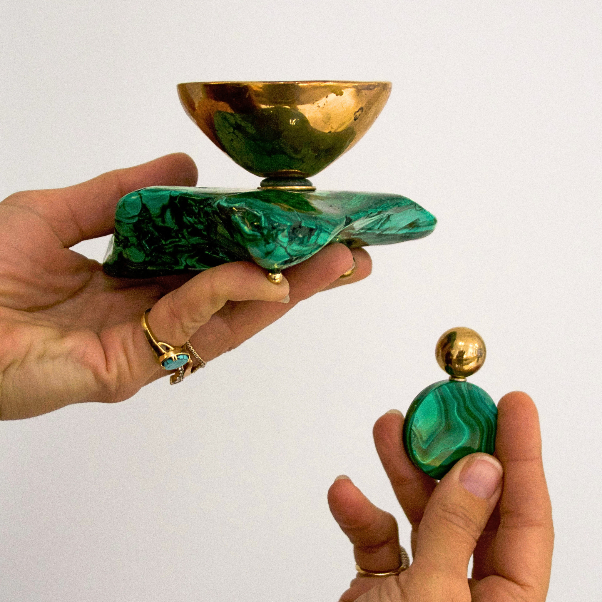 Two hands holding a green malachite and bronze decorative object and bell against a white background 