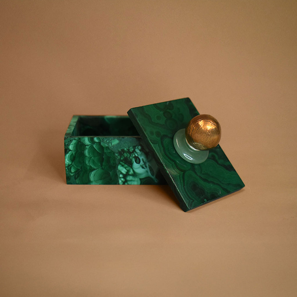 Malachite box with an open lid composed of Aventurine gemstone and bronze ball on top of lid on a beige background