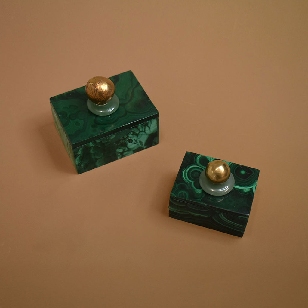 Two malachite boxes with malachite lids topped with aventurine gemstones and bronze balls on a beige background