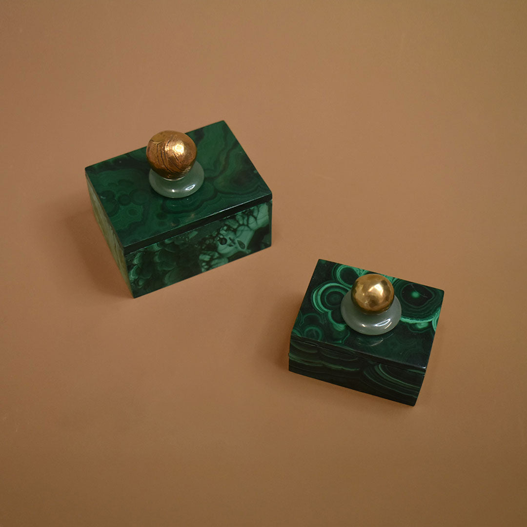 Two malachite boxes with malachite lids topped with aventurine gemstones and bronze balls on a beige background