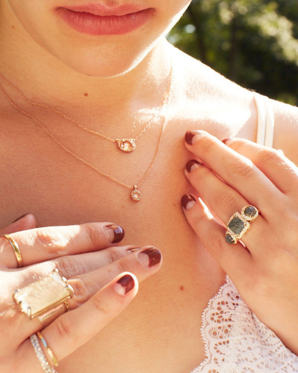 A close-up lifestyle shot of a woman wearing layered gold necklaces and multiple gold rings, including one with a green rectangular stone and another with a large flat gold plate, against a warm, sunlit outdoor background.