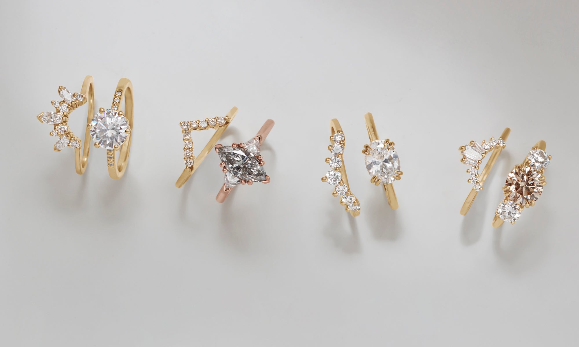 A cascade of different silhouettes of engagement rings with differently shaped center diamonds stacked with white diamond bands on a grey background.