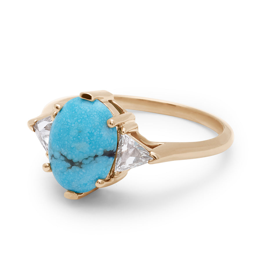 Angled view of a three stone engagement ring with an opal shape turquoise center stone with two white diamond trillion side stones set in yellow gold.