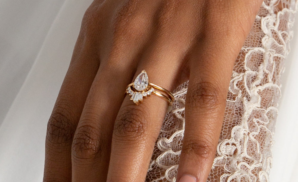 A medium complextion model wears a white diamond pear shaped engagement ring stacked on top of a baguette band over a white lace detail.