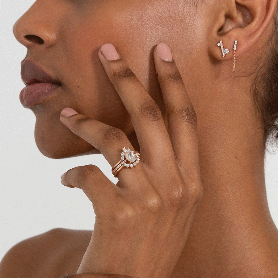 A single earring with a straight line of white diamond pave with white diamond accent stones on either side set in a yellow gold on a darker-complextion model's ear with a stack of three rings on her ring finger.