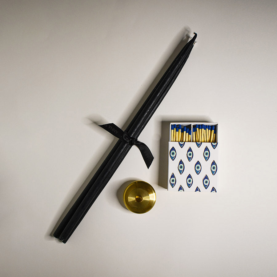 A brass candle holder with a three pack of tall black candlesticks and a pack of matches in evil eye packaging next to it.