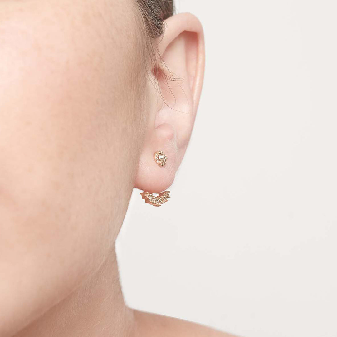A single tiny champagne diamond center stone earrings with a halo of champagne diamond set in yellow gold on a light complexion ear.
