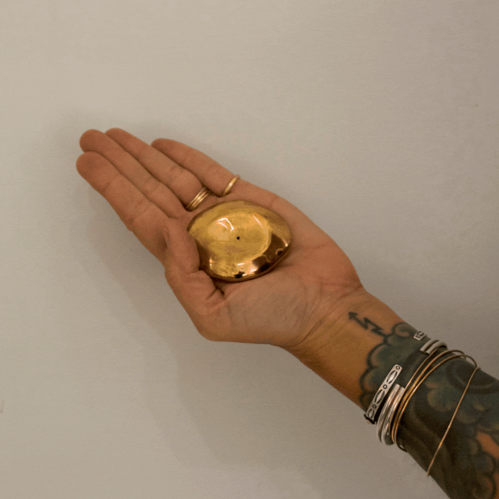 A gif of a hand holding a golden bronze small orb incense holder and tapping a blue incense stick into it against a white background 