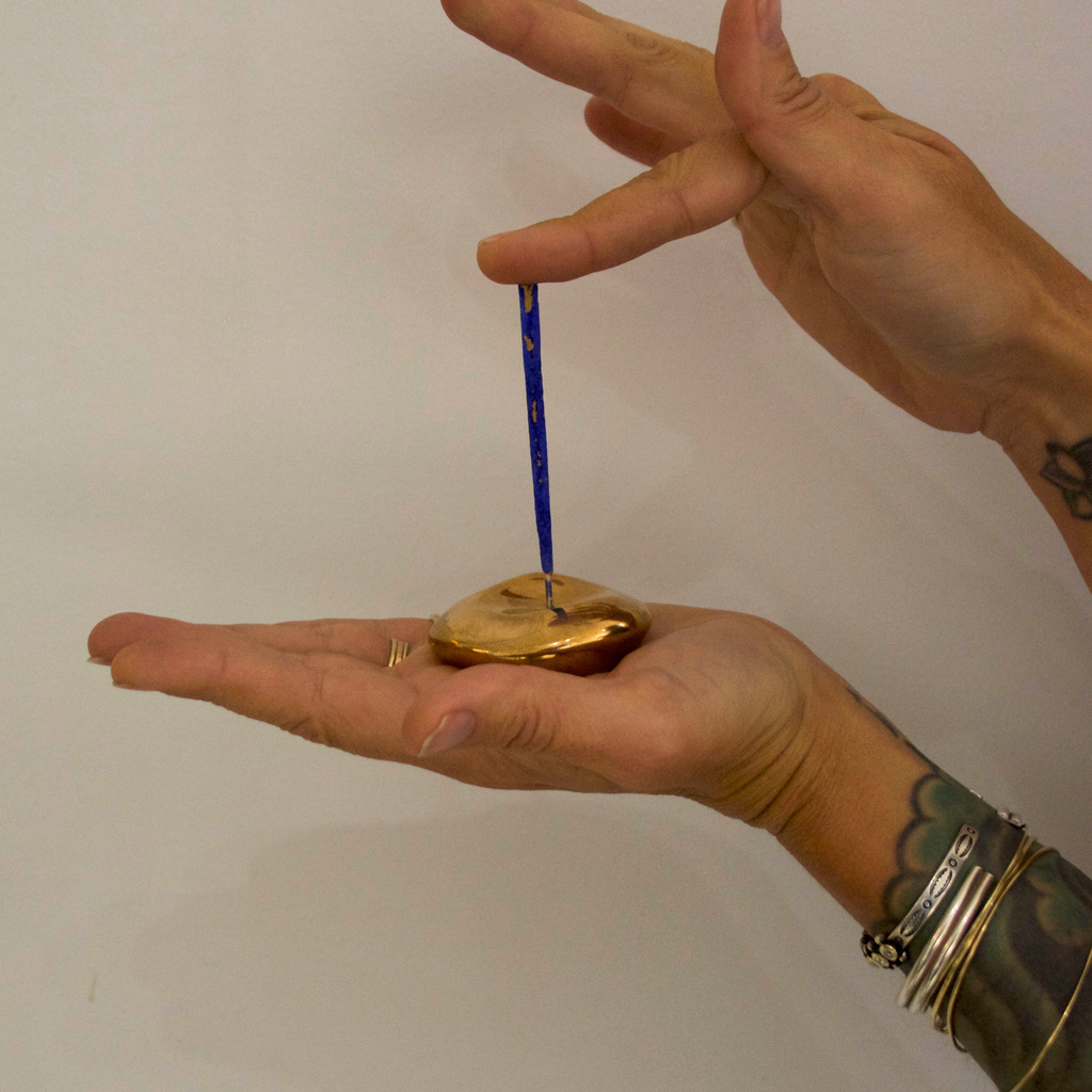 A hand holding a golden bronze small orb incense holder and tapping a blue incense stick into it against a white background 