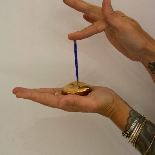 A hand holding a golden bronze small orb incense holder and tapping a blue incense stick into it against a white background 