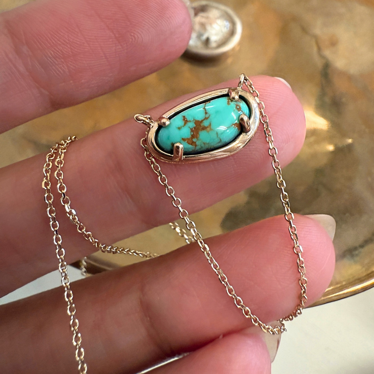 A gold chain necklace with an oval turquoise stone pendant held on a person's fingers. The pendant features a vibrant turquoise center with brown matrix veining, set in a gold bezel.