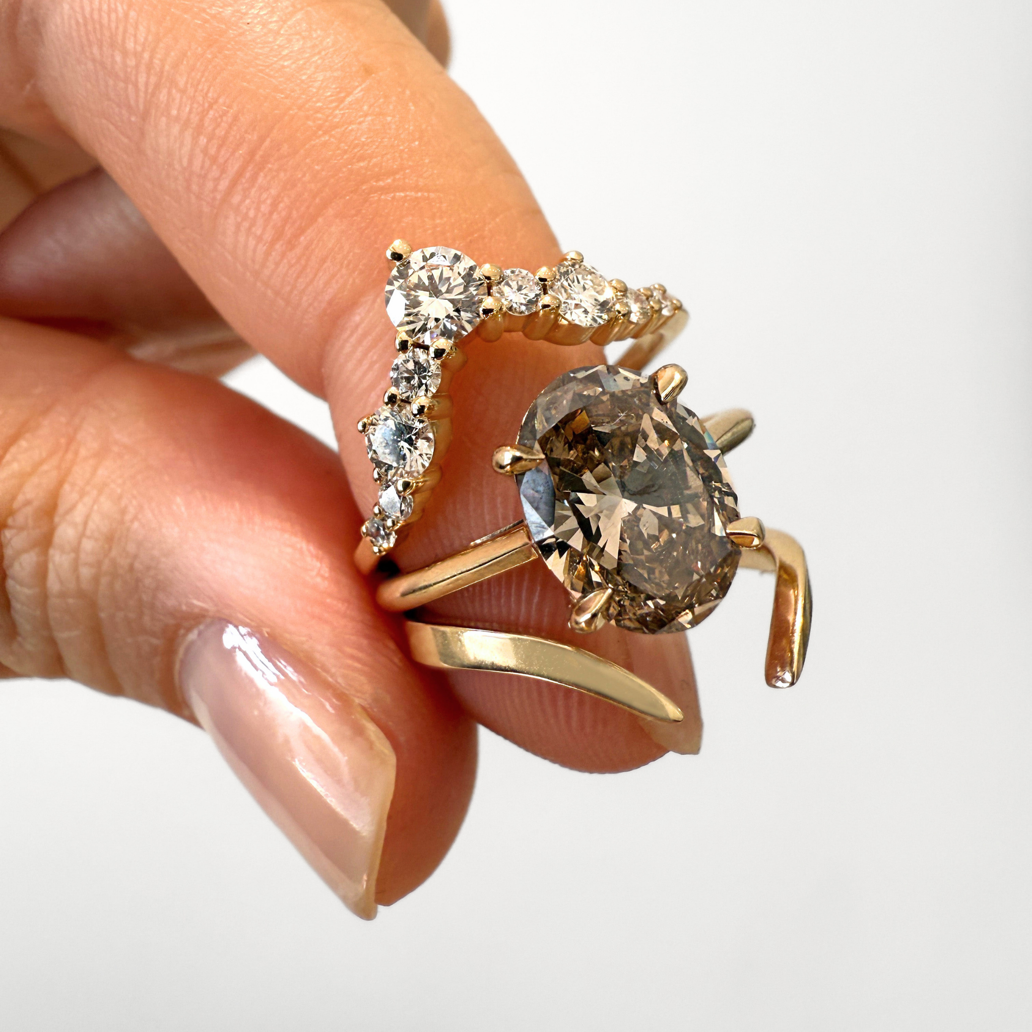 A close-up of a hand holding two rings against a plain light background. The larger ring features a prominent oval smoky-gray center stone with a gold band, while the smaller ring has a curved row of clear stones on a gold band.