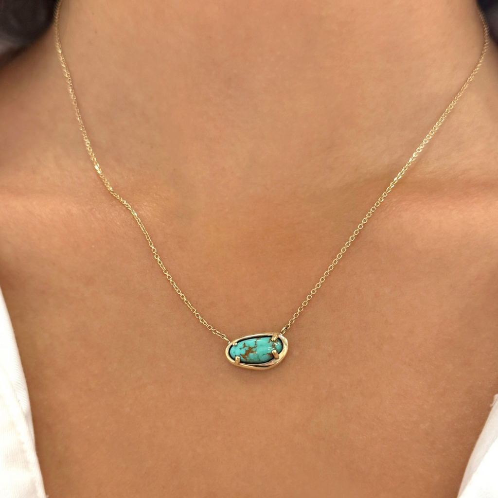 A close-up of a gold chain necklace worn on a model's neck, featuring an oval pendant with a turquoise stone set in a gold frame.
