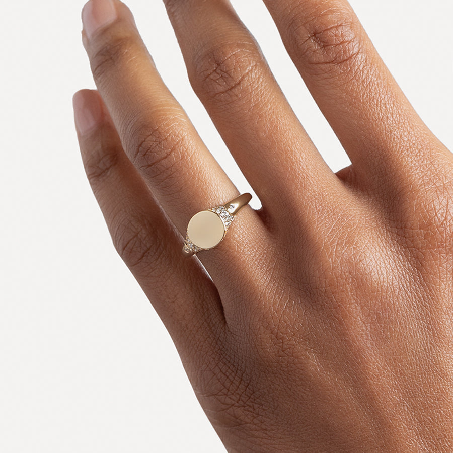 A solid gold signet ring with two white diamond accents on either side and a pave set around the top of the ring and set in yellow gold on a medium-complextion ring finger.