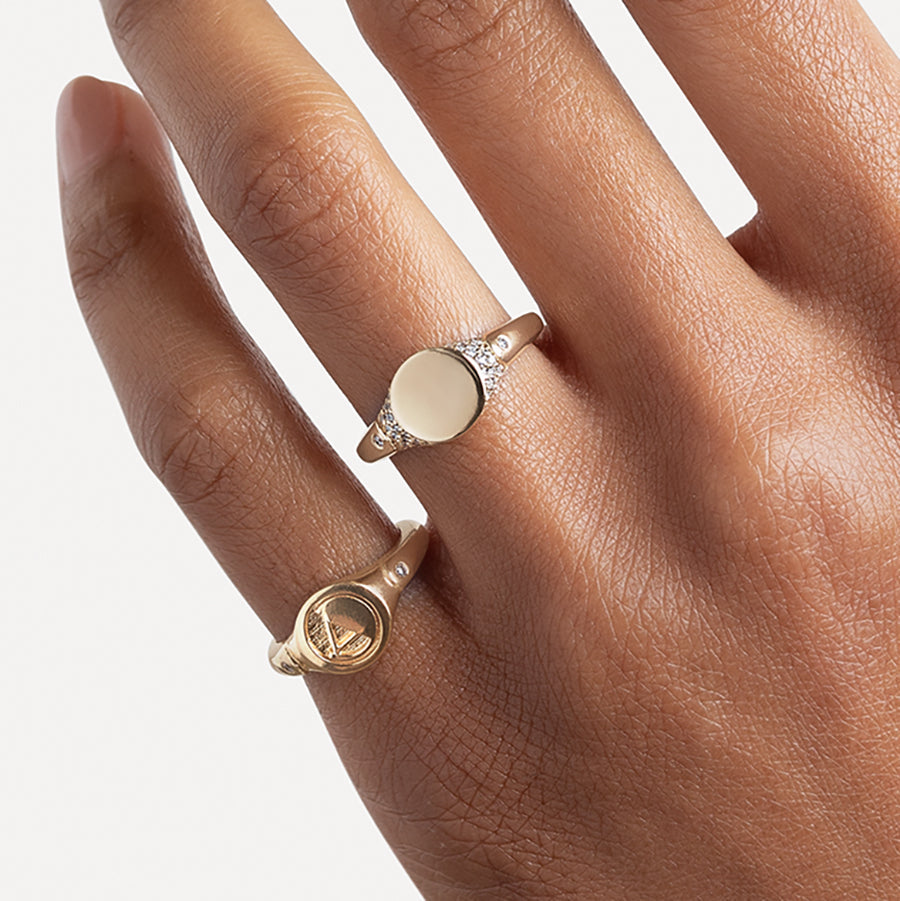 A solid gold signet ring with two white diamond accents on either side and a pave set around the top of the ring and set in yellow gold on a medium-complextion ring finger and a monogrammed signet ring with an "A" on the pinky finger.