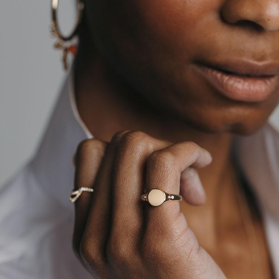 A solid gold signet ring with two white diamond accents on either side and set in yellow gold on a darker-complextion pinky ring with a diamond accent band on her pointer finger.