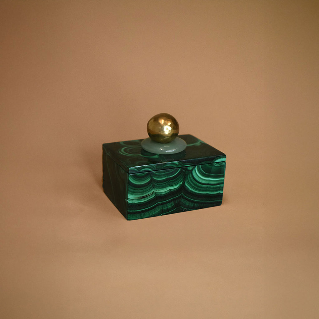 Small malachite box with an Aventurine gemstone and bronze ball on top of lid on a beige background