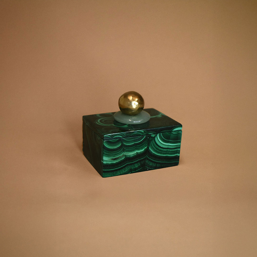 Small malachite box with an Aventurine gemstone and bronze ball on top of lid on a beige background