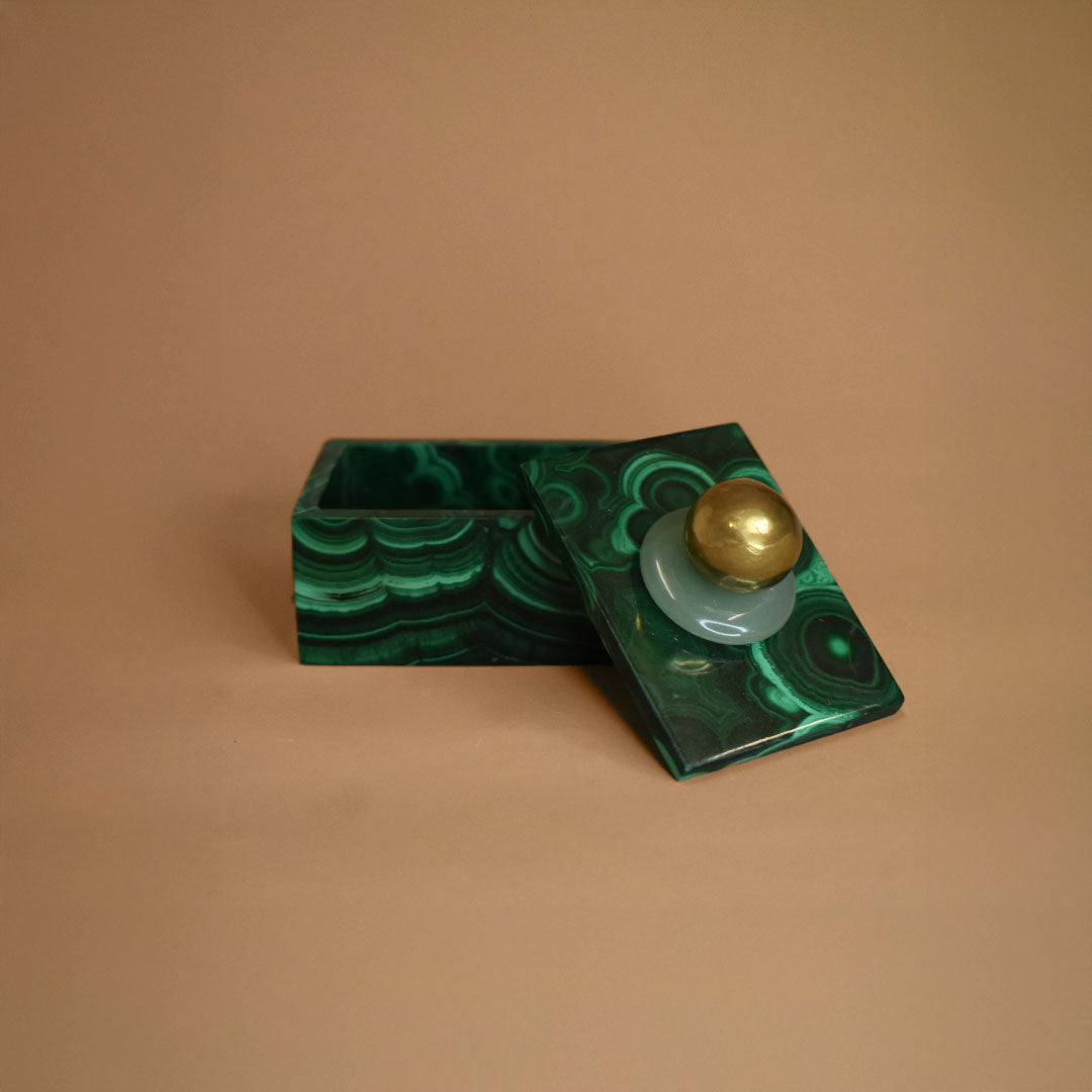 Small malachite box open with an Aventurine gemstone and bronze ball on top of lid on a beige background