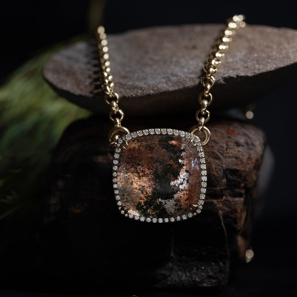 A cushion shaped quartz charm with a desert like scene in a bezel setting with claw prongs and a white diamond halo set in yellow gold on a yellow gold chain in a dark setting over a few rocks.