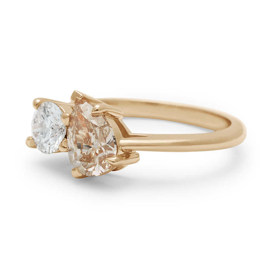 Angled view of a two diamond ring with a pear champagne diamond set next to a round white diamond set in yellow gold.