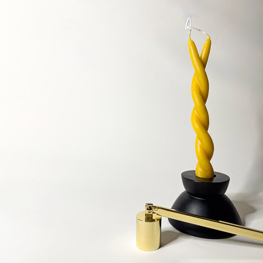 A black candle holder holding a yellow twisted candle to symbolize a twin flame with a brass candle snuffer next to it.