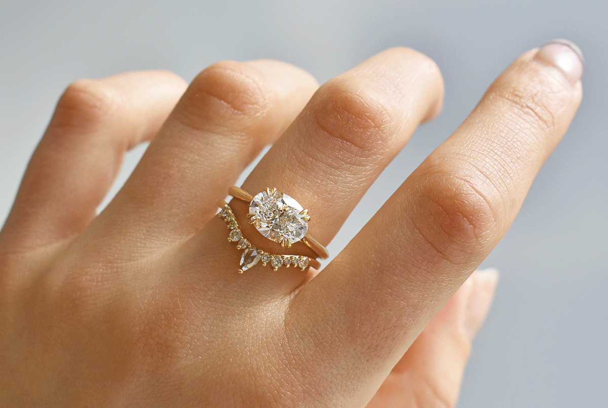 A hand wears a Bea Solitaire ring with a white diamond set east-west paired with a curved white diamond wedding band.