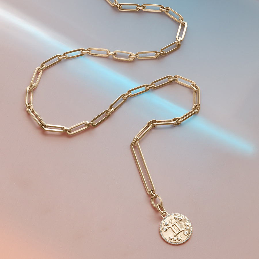 Mixed link chain necklace in the center with a Gemini constellation charm at the end set on a blue/purple/pink background.