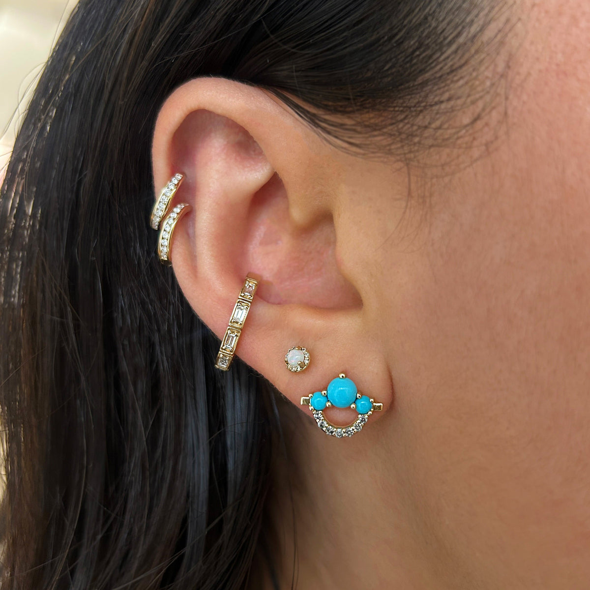 Close-up of a person's ear showcasing a multi-piece ear cuff and stud jewelry set. The set includes two gold huggie hoop earrings with clear pavé stones, a gold cartilage hoop, a small gold stud, and a gold crescent-shaped stud featuring a vibrant turquoise blue center surrounded by clear pavé stones.