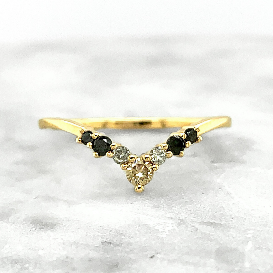 A v-shaped wedding band featuring round diamonds of different colors and shapes, including one largest champagne diamond in the center followed by small grey diamonds and finally followed by two black diamonds on either side, set in yellow gold. 