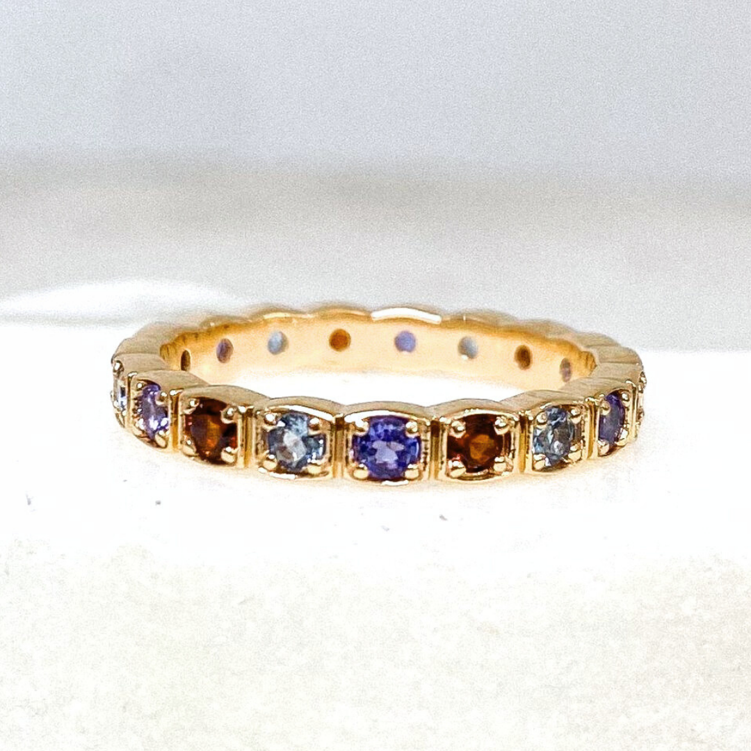 A gemstone eternity band featuring alternating red, blue, and purple gemstones set in yellow gold against a white background. 
