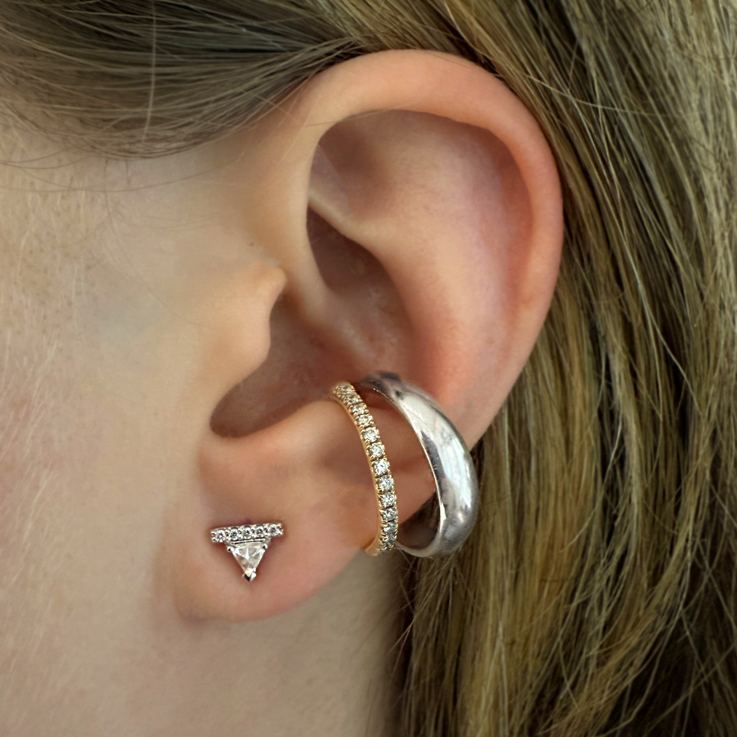 An ear featuring one single white triangle diamond stud earring and a double hoop ear cuff that features a yellow gold hoop with white diamonds and a silver hoop