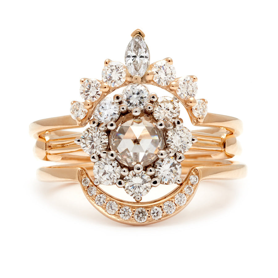 A cluster engagement ring with a rose cut champagne diamond center stone and round white diamond stones surrounding it set in yellow gold stacked between a white diamond marquise nesting band and a curve white diamond nesting band.