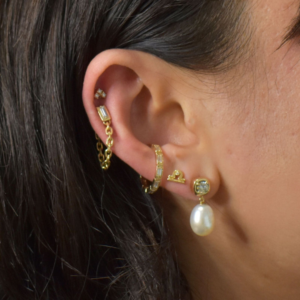 An ear featuring multiple earrings including a white diamond stud earring, a baguette diamond earring with a yellow gold chain, a baguette diamond gold ear cuff, a yellow gold libra zodiac stud, and a drop earring with champagne diamond with a pearl droplet. 