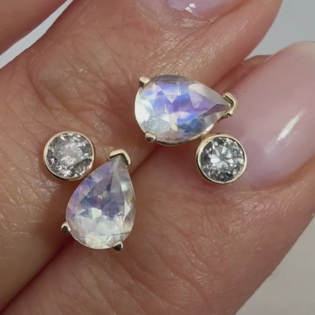 A video of two toi et moi stud earrings featuring a pear cut moonstone and a round brilliant grey diamond in between two fingers 