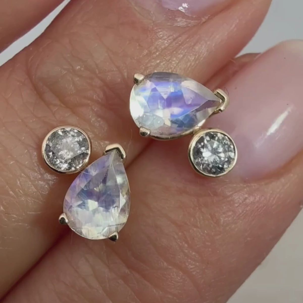 A video of two toi et moi stud earrings featuring a pear cut moonstone and a round brilliant grey diamond in between two fingers 