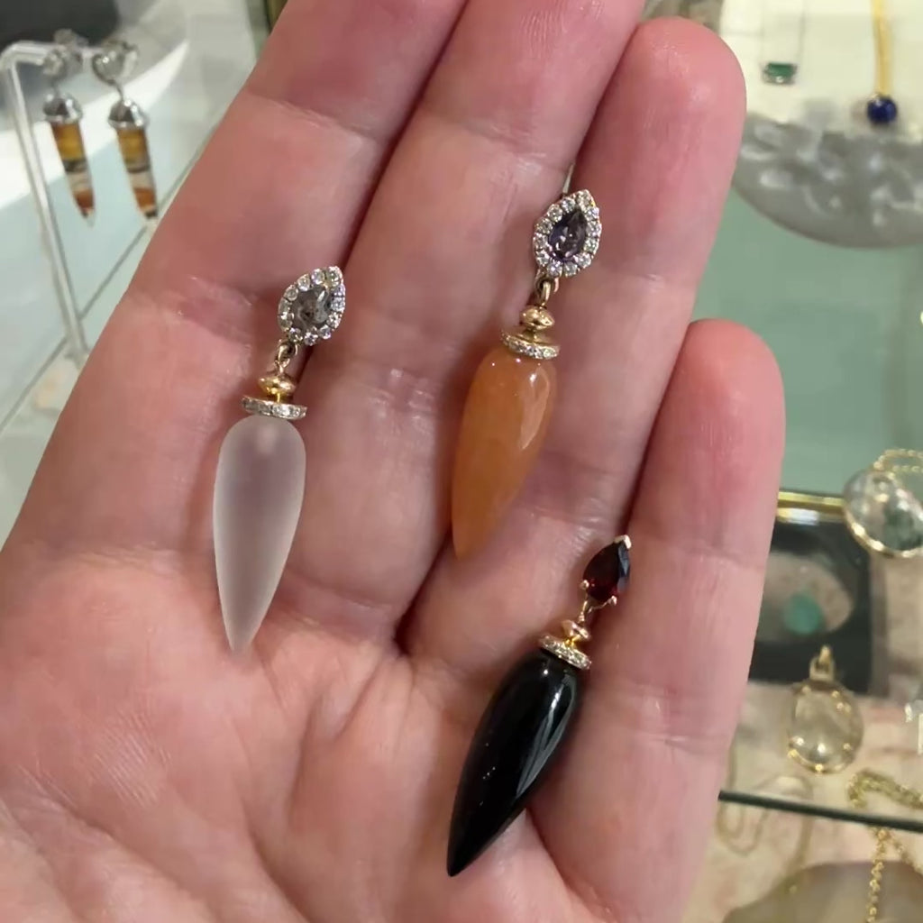 A video of three gemstone drop earrings with clear, orange, and black gemstones in between the fingers of someone's hand