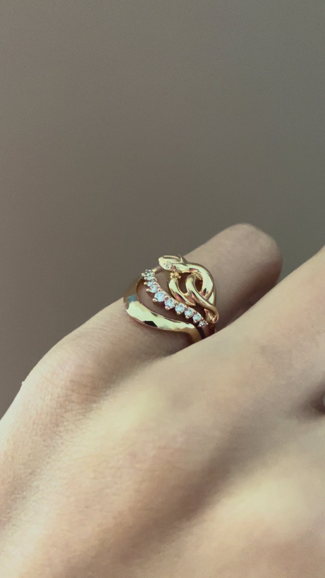 A video of a woman's pinky finger wearing a yellow gold serpent ring stacked with white diamond and yellow gold bands against a beige background 