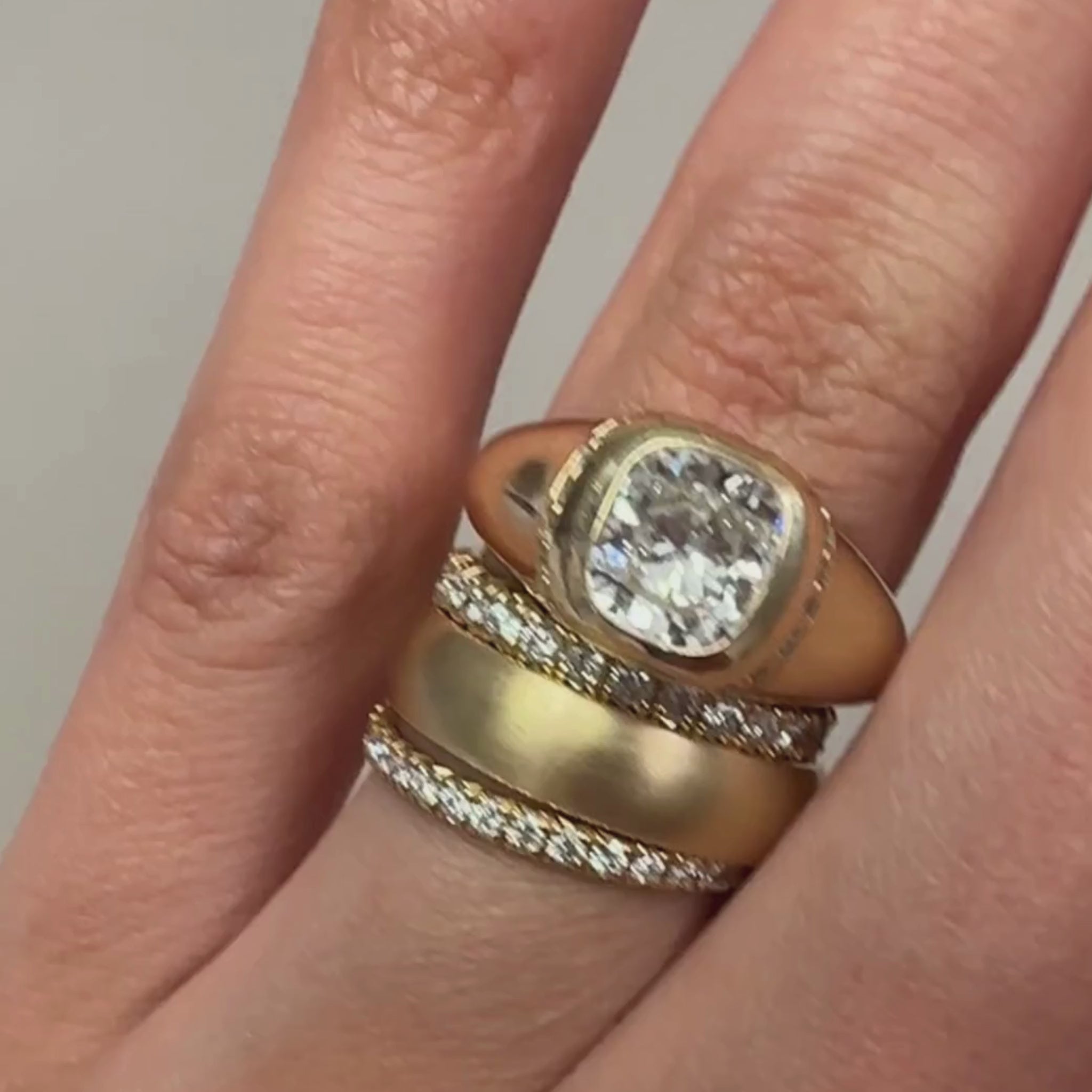A video of a chunky gold white diamond ring and two white diamond band and one satin-finish yellow gold band on a woman's ring finger against a greyish-beige background