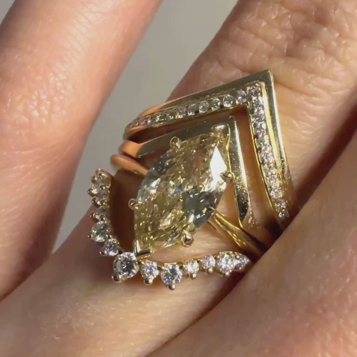 A woman's ring finger wearing multiple yellow gold and diamond rings including a big yellow marquise diamond and white diamond bands against a grey background