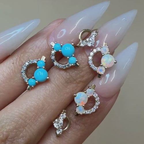 A video of opal, blue turquoise, and diamond gemstone earrings set in yellow gold in between the fingers of a woman's hand. She has long, white, pointed almond shaped nails. 
