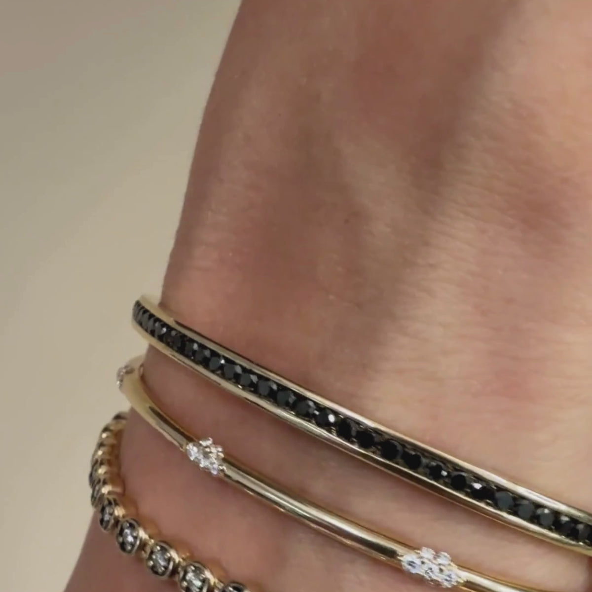 A video of three yellow gold and black diamond and white diamond bracelets on a woman's rotating wrist against a beige background