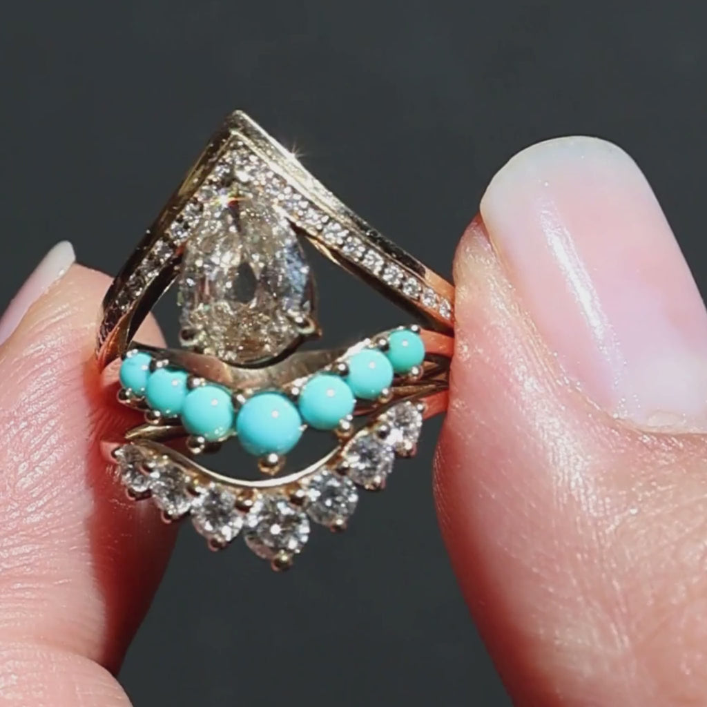 A video of three rings held in between a woman's fingers sparkling in the sunlight against a black background. The rings feature one pear cut champagne diamond yellow gold engagemnet ring, a turquoise cabochon yellow gold curved band, and a white diamond yellow gold curved band.