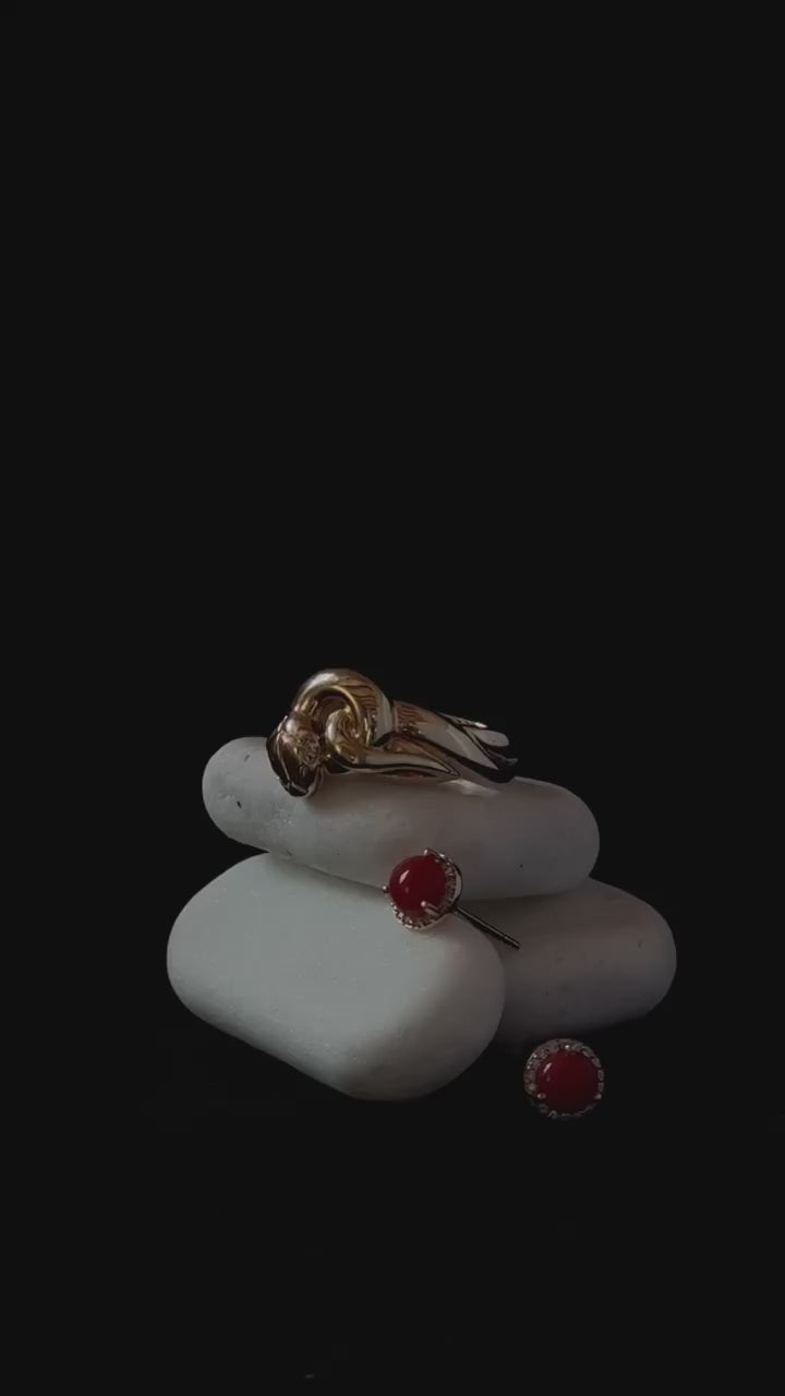 A video of three white rocks against a black background. On the rocks there is styled a yellow gold serpent ring, and two red coral stud earrings featuring diamond halos 