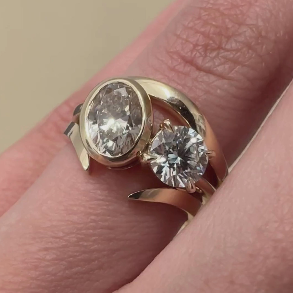 A close-up of a woman's ring finger wearing a toi et moi champagne diamond and white diamond yellow gold engagement ring and two yellow gold bands against a beige background